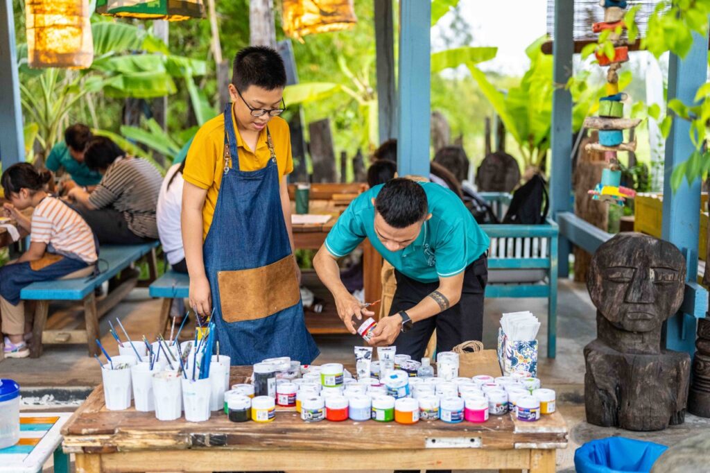 Alt text: Young boy learning wood carving and painting at a workshop at Lang Cui Lu - Driftwood Village in Hoi An, Vietnam, guided by a master craftsman.