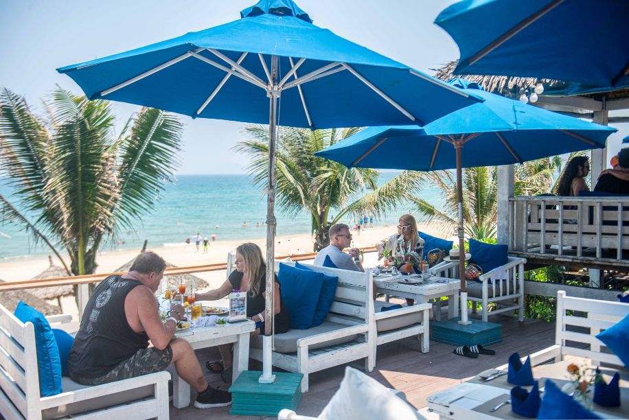 Guests relaxing under blue beach umbrellas at Shore Club, An Bang Beach, toasting drinks and enjoying the Easter Sunday atmosphere.